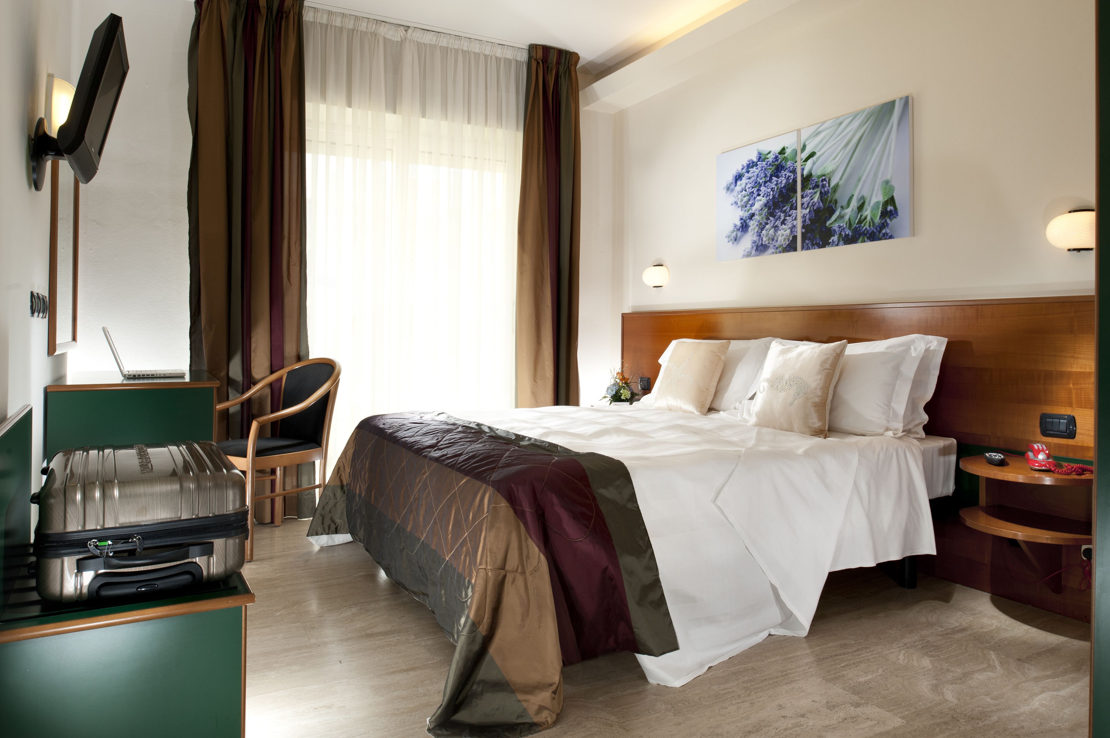 Habitación de hotel con cama, escritorio con maleta, una silla, televisión montada en la pared y un cuadro.