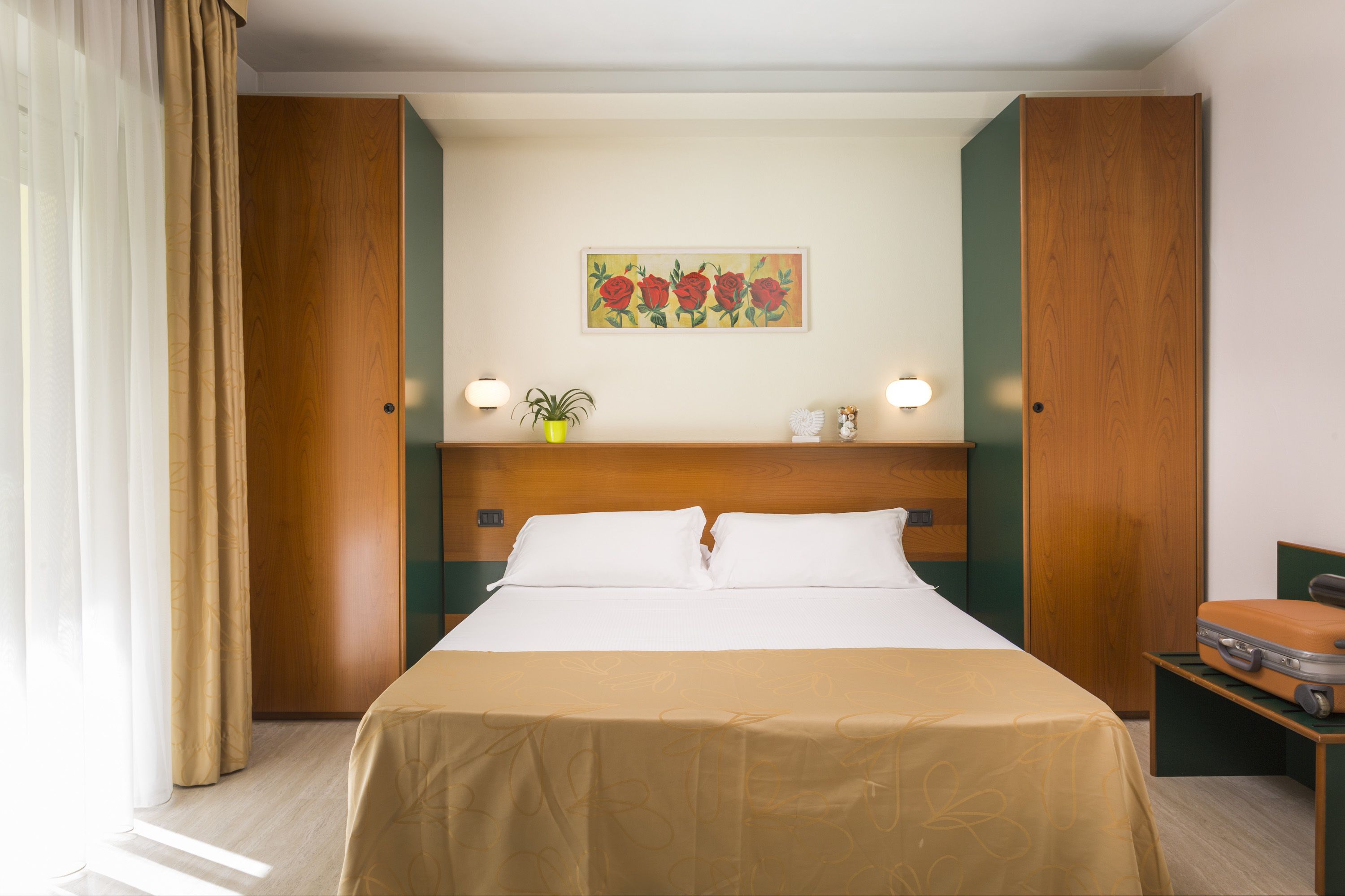 Ein Hotelzimmer mit einem Bett, einem Bild an der Wand, einer Zimmerpflanze und einem Koffer auf einem Beistelltisch.