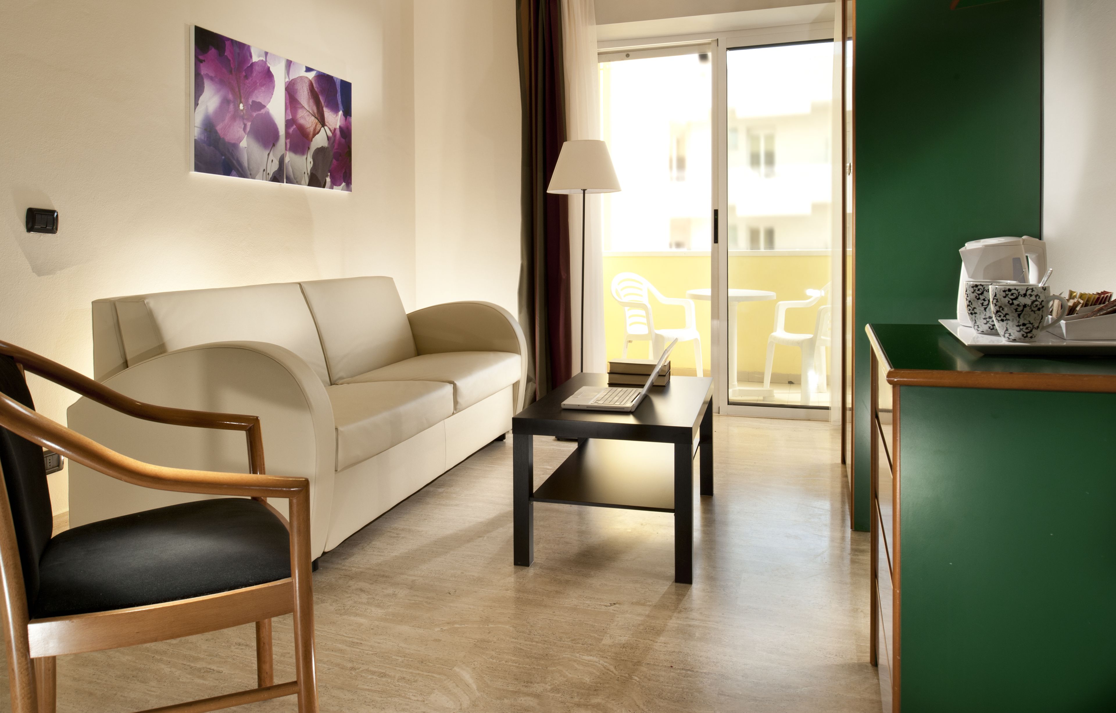 A modern living room with a sofa, a wooden chair, a small table with a laptop, a green cabinet, and a balcony with a chair and table.