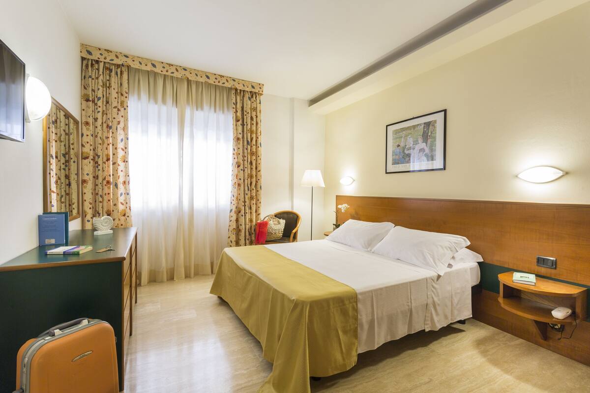 A hotel room with a bed, desk, chair, and a suitcase.