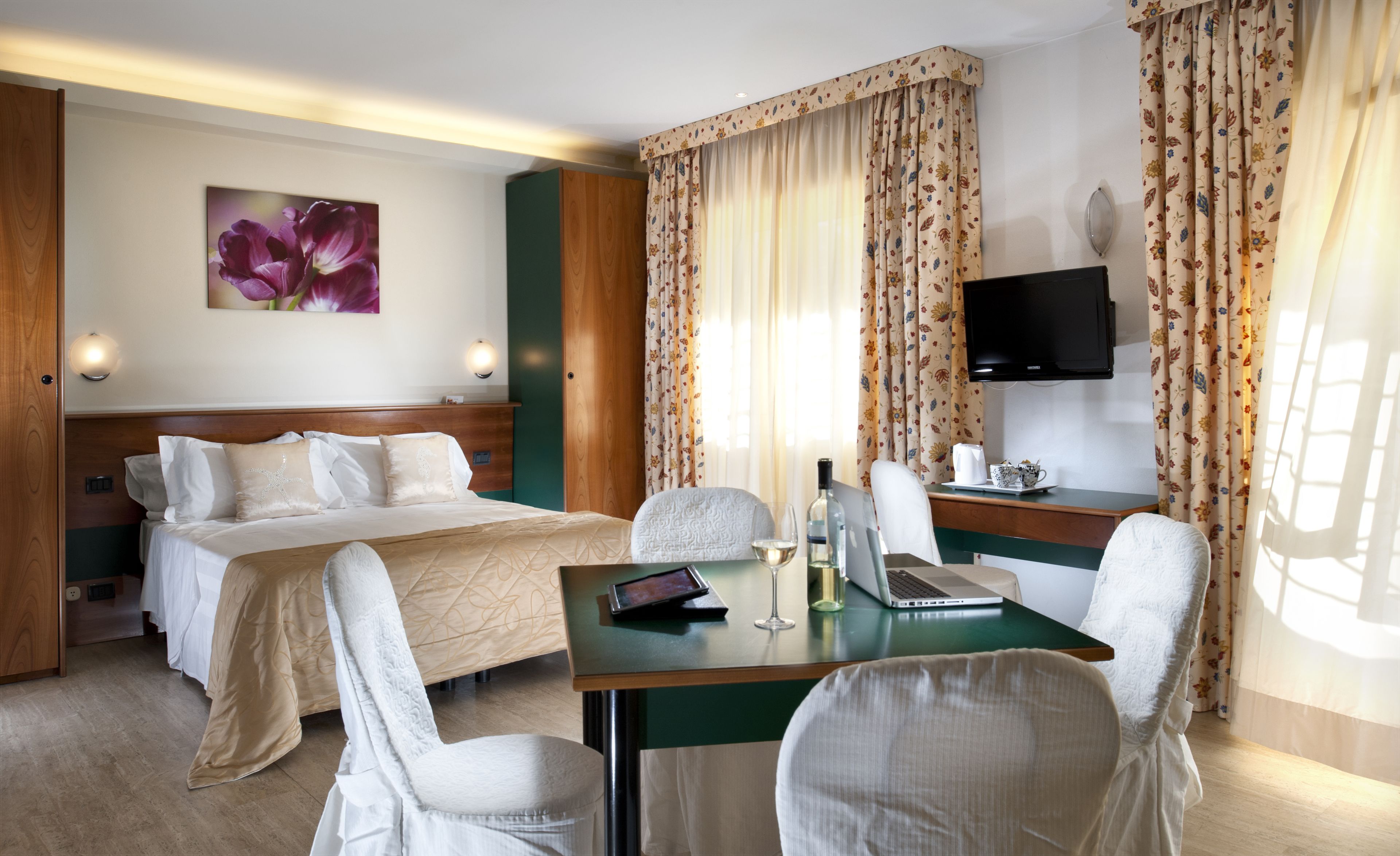 A hotel room with a bed, two chairs, a desk with a laptop, a TV, and a floral curtain.