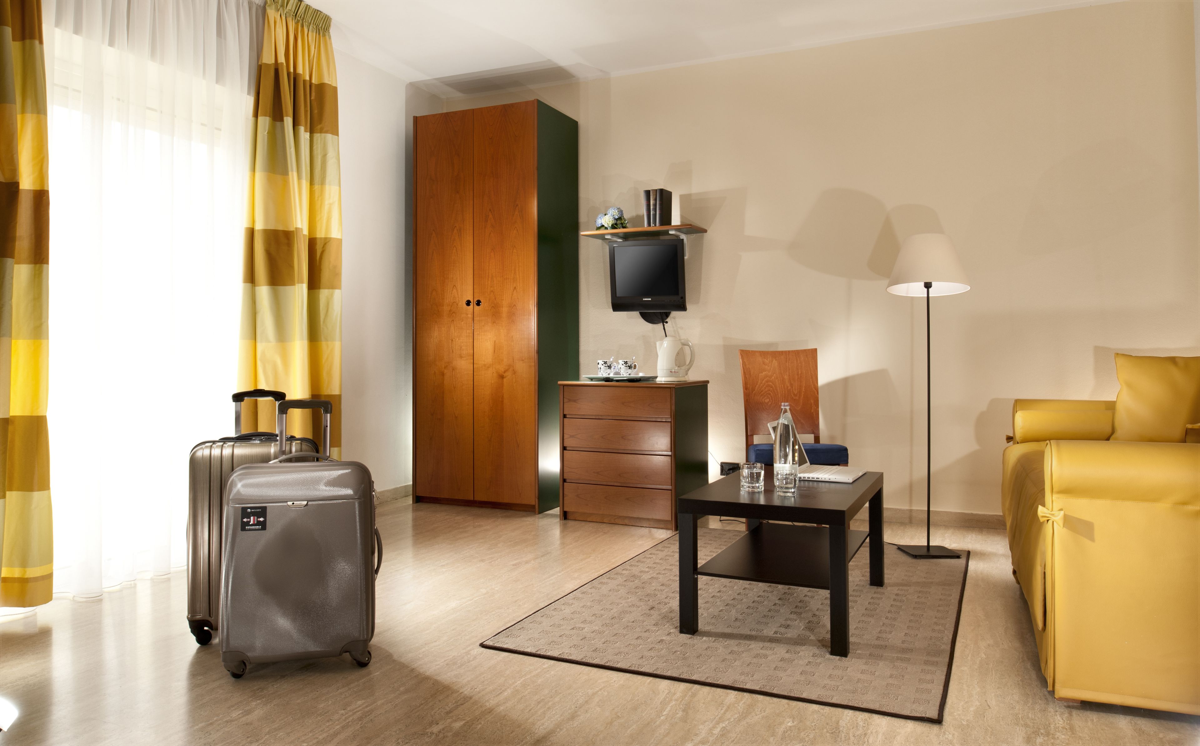 Ein Hotelzimmer mit Holzschrank, Schreibtisch mit Lampe, gelbes Sofa und Koffer.