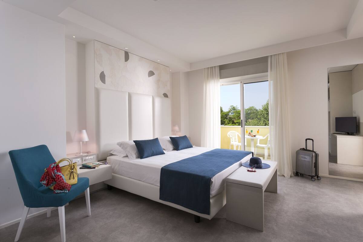 A modern hotel room with a bed, a desk, a chair, a TV, and a balcony with a view.