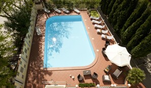 Vista aérea de una piscina con tumbonas y sombrillas, rodeada de vegetación y un solárium empedrado.