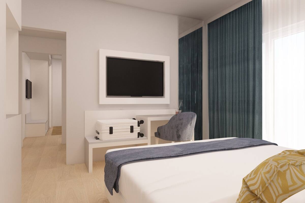 A modern hotel room with a large bed, a flat-screen TV mounted on the wall, a desk with a suitcase, and a chair.