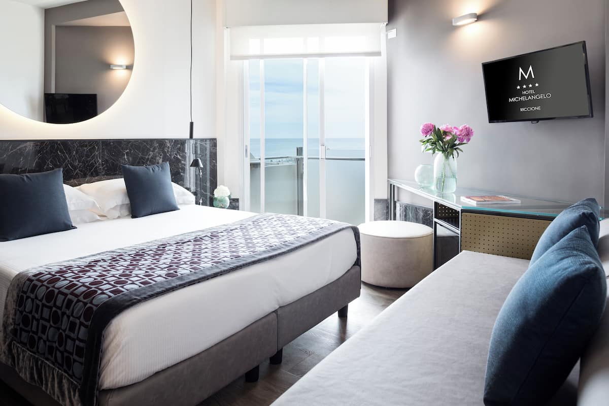 A modern hotel room with a large bed, a sofa, a TV, and a balcony with a view of the sea.