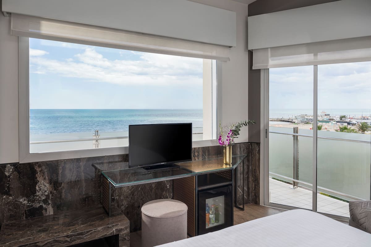 A modern hotel room with a large window offering a view of the sea and a balcony with a glass railing.