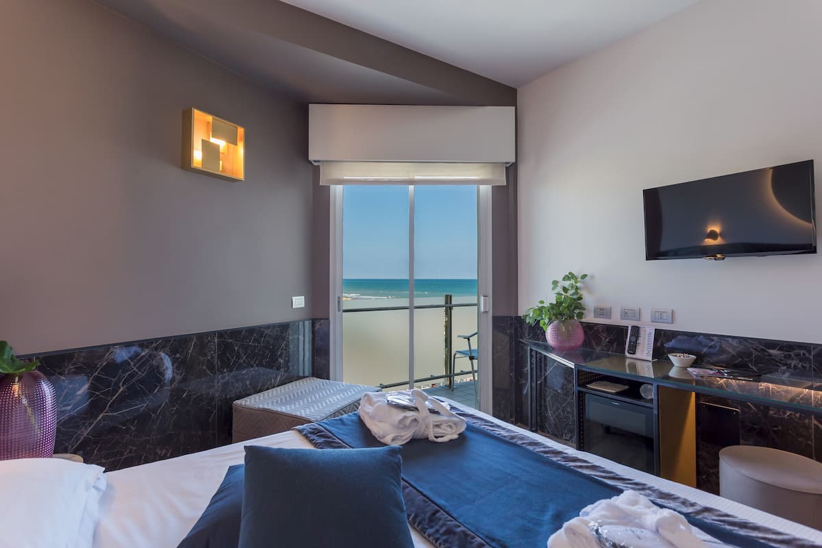 A hotel room with a bed, a desk, a TV, and a view of the beach.