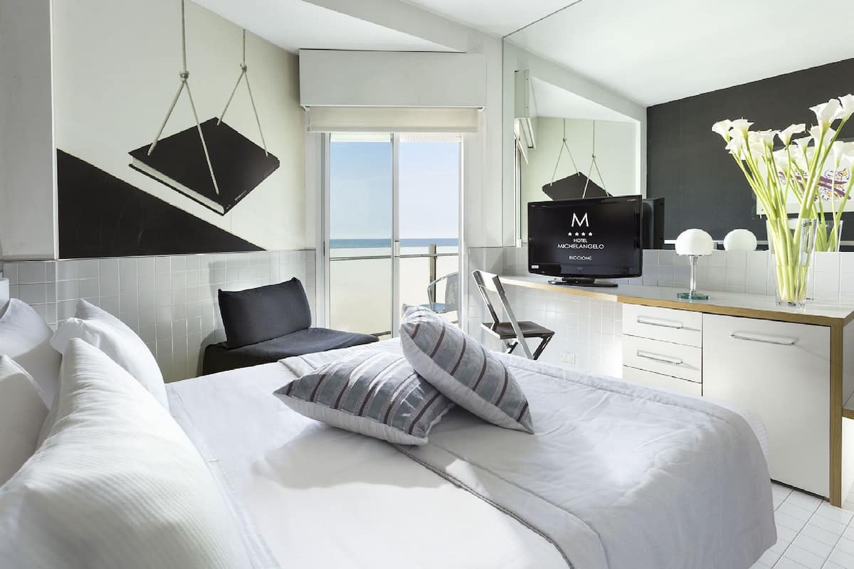 A modern hotel room with a bed, a desk, a television, and a view of the sea.