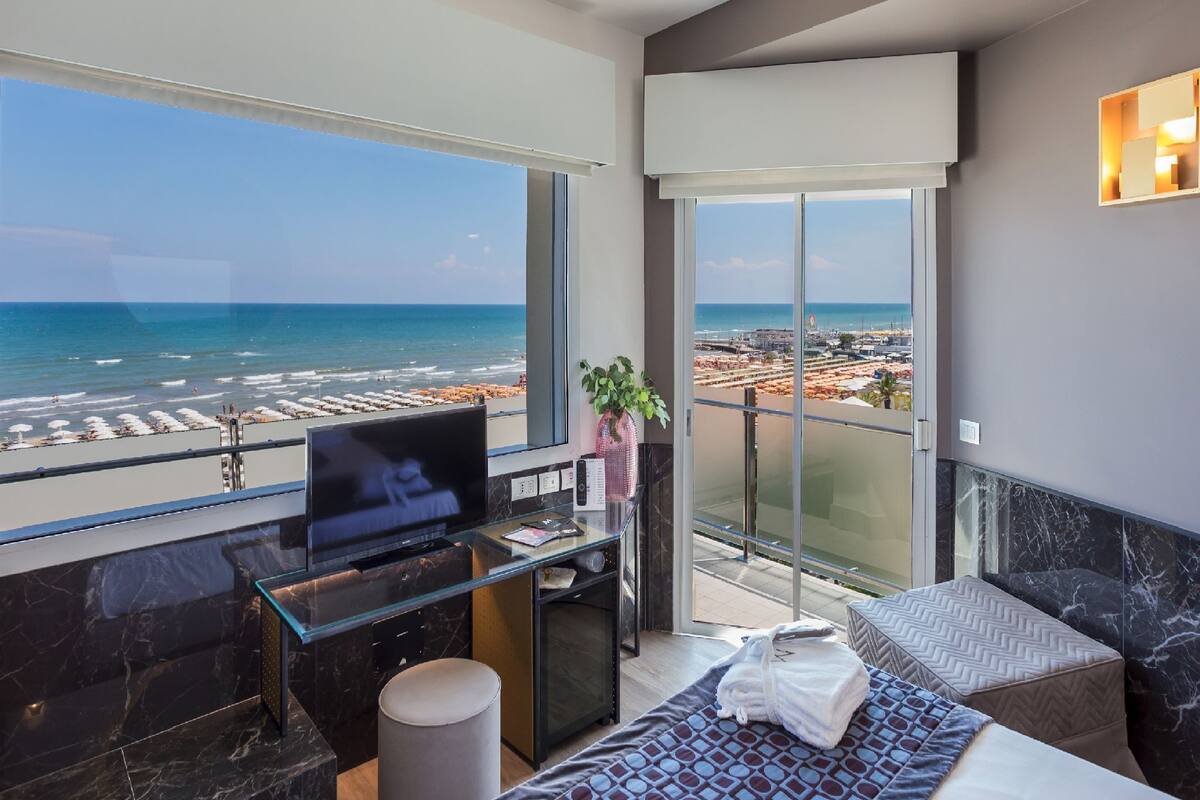 A modern hotel room with a balcony overlooking a beach, a TV, and a glass desk.
