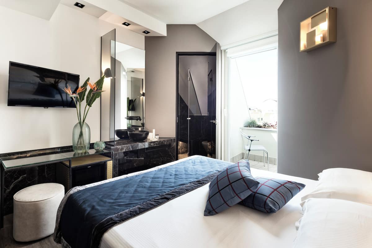 A modern hotel room with a large bed, a flat-screen TV mounted on the wall, a vanity with a mirror, and a view of the city through the window.