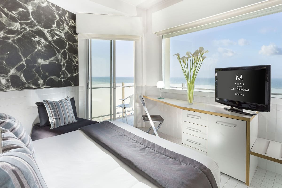 A modern hotel room with a large bed, a desk with a TV, a balcony with a view of the sea, and a marble wall.