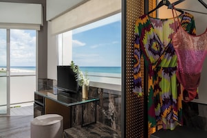 A modern room with a large window overlooking a beach, a glass table, and a colorful dress hanging on the wall.