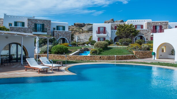 A pool area with lounge chairs, a stone wall, and a view of buildings with red shutters and balconies. - Naxos Palace Hotel (Naxos)