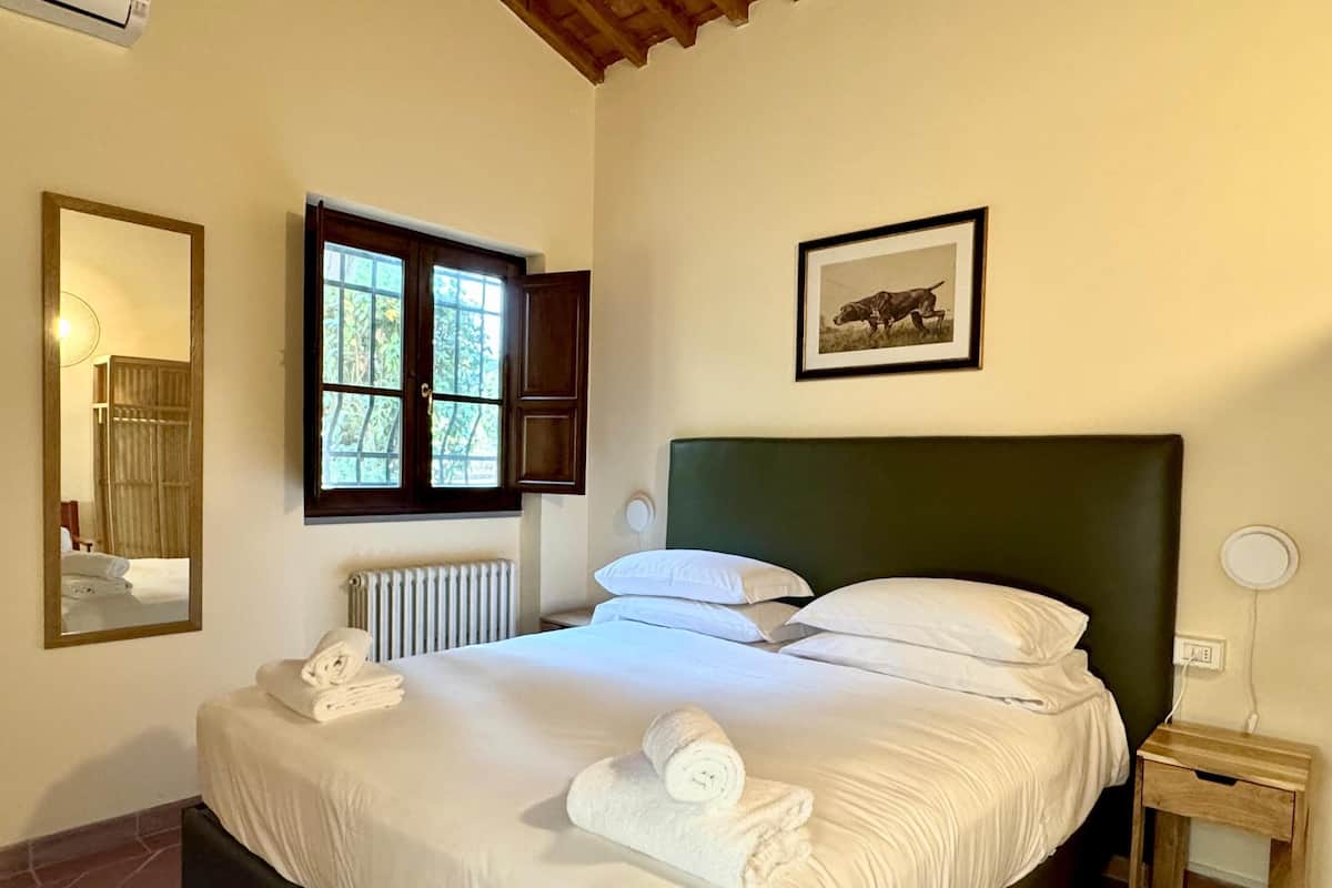 A neatly made bed with white linens and towels, a window with wooden shutters, a framed picture on the wall, and a small bedside table.