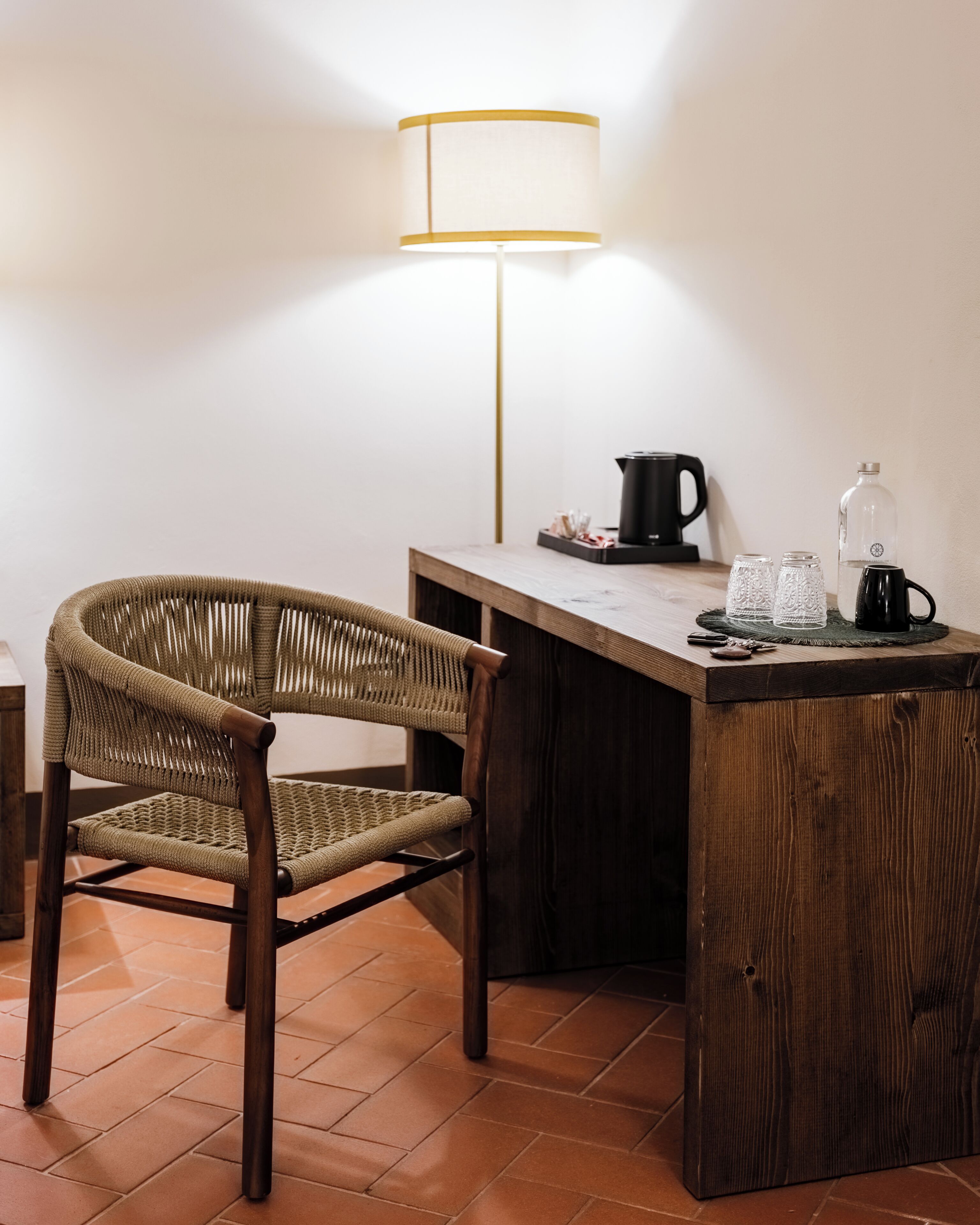 Une pièce avec un bureau en bois, une chaise en osier, une lampe, une machine à café et un bocal en verre.