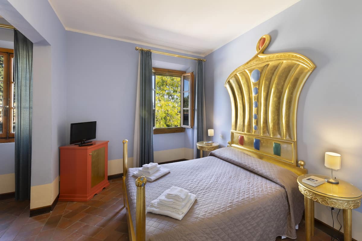 A hotel room with a bed, a television on a cabinet, a window with curtains, a bedside table with a lamp, and a decorative mirror on the wall.