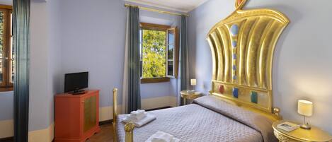 Habitación de hotel con cama, televisor en un mueble, ventana con cortinas, mesita de noche con lámpara y un espejo decorativo en la pared.