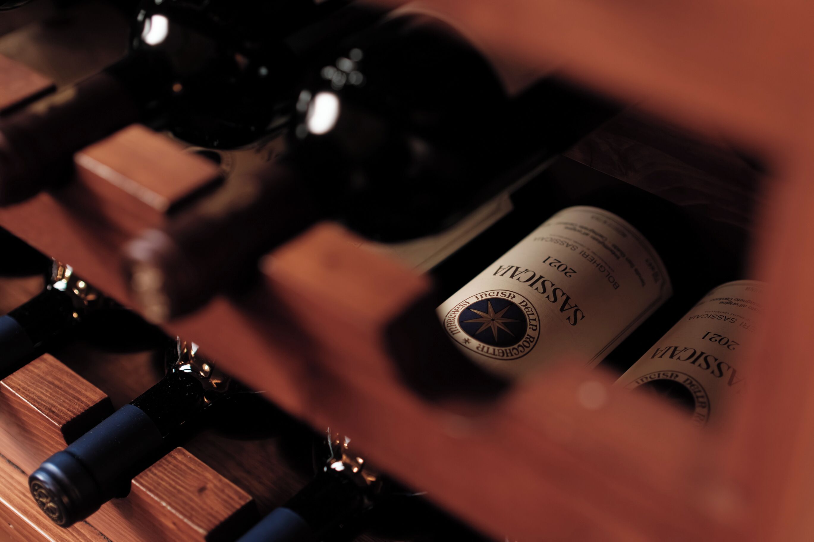 Close-up de garrafas de vinho com etiquetas, uma superfície de madeira e um objeto desfocado em primeiro plano.