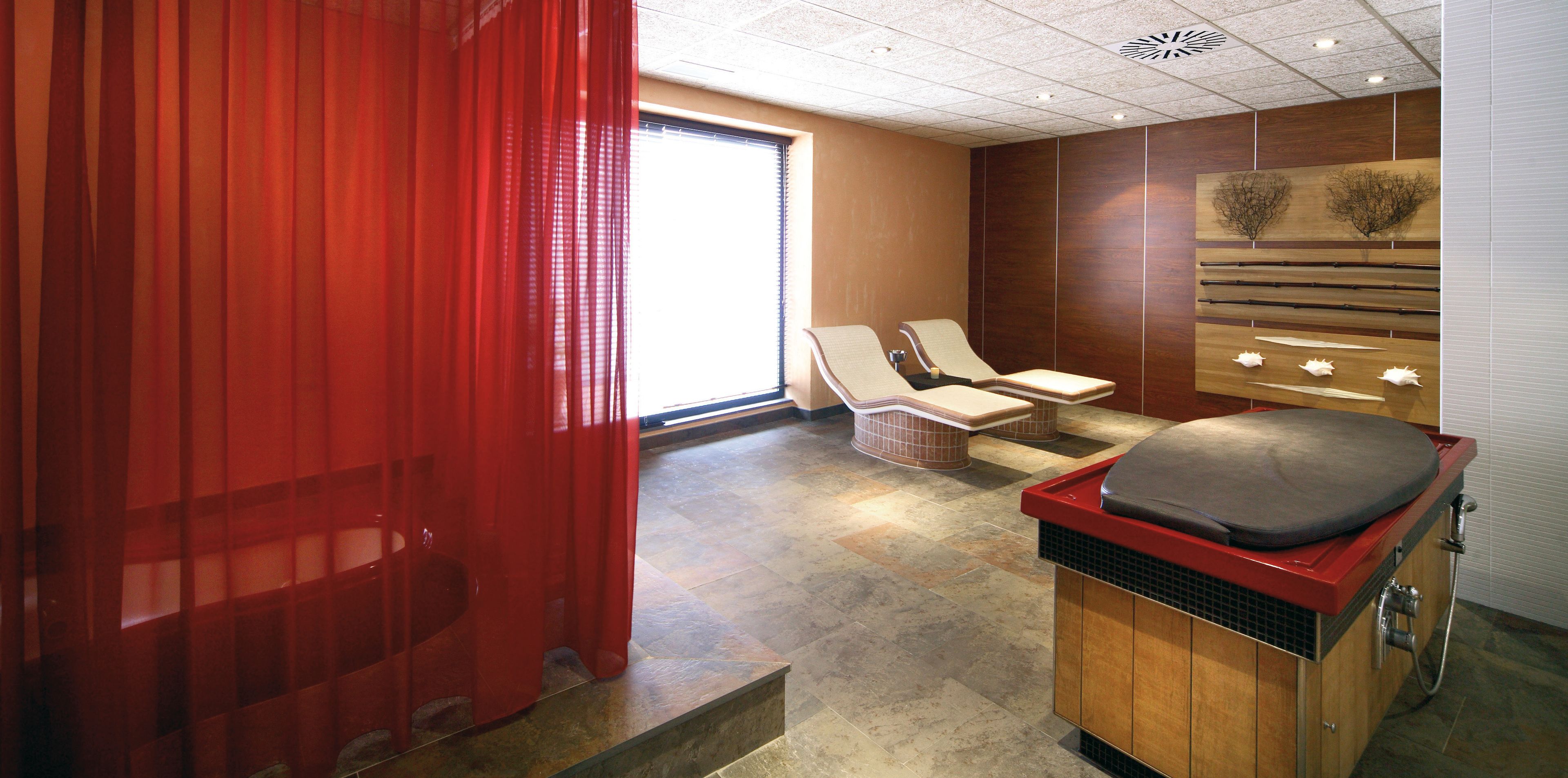 Une salle de spa avec un rideau rouge, deux fauteuils, une table de massage et un panneau en bois fixé au mur, orné d’une œuvre d’art abstraite.
