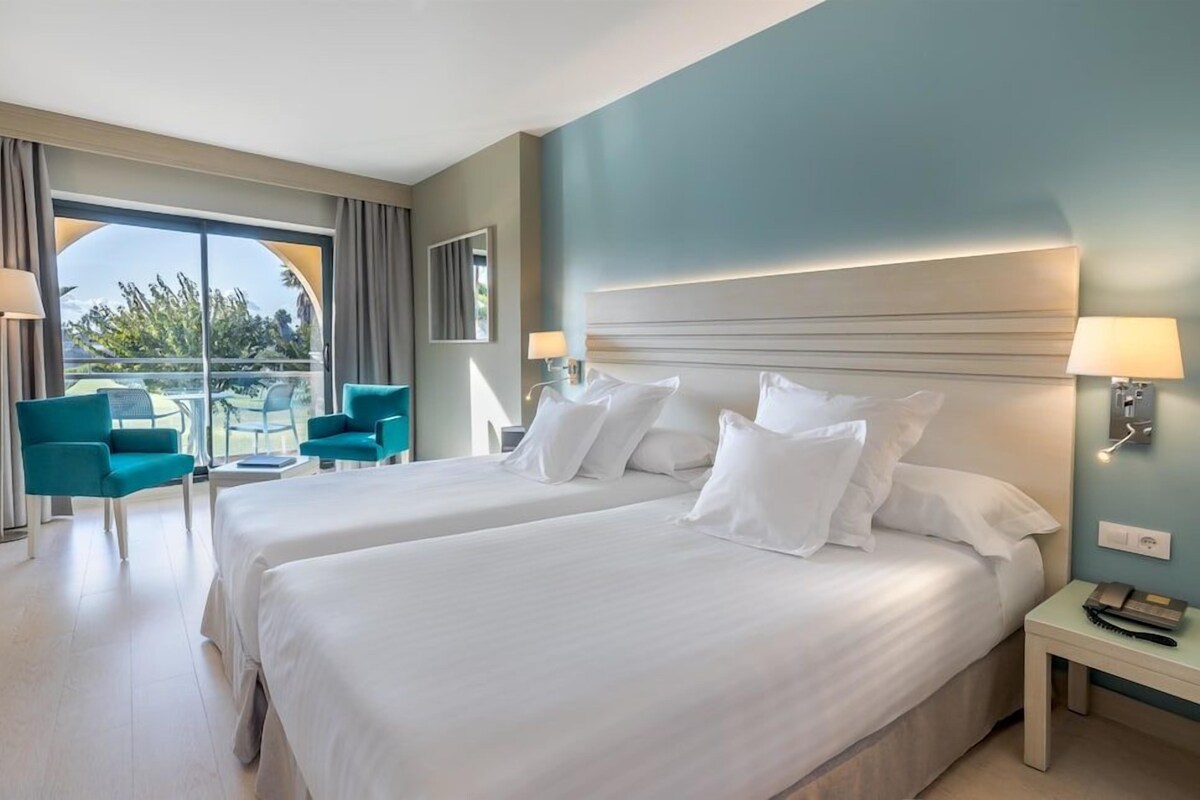 A hotel room with two beds, a balcony with chairs, and a view of greenery.