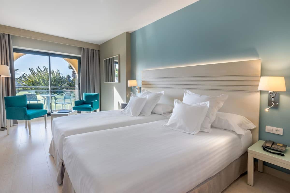 A hotel room with two beds, a balcony, a teal armchair, and a nightstand with a lamp.