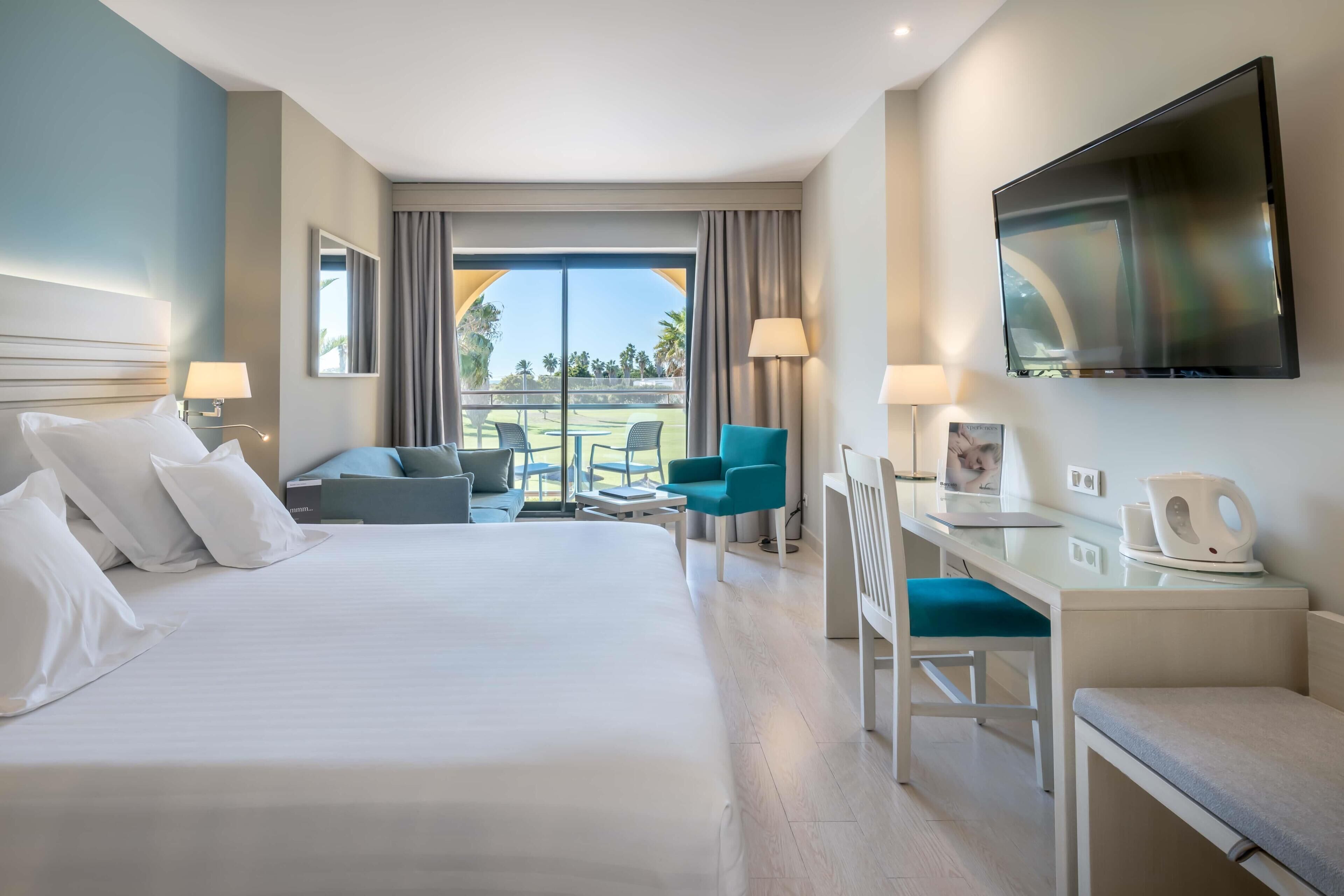 A hotel room with a bed, a desk, a chair, a TV, and a balcony with outdoor seating.