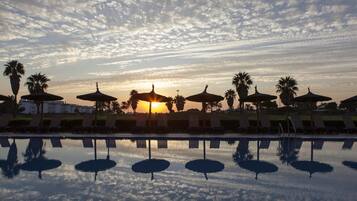 Een zwembad met ligbedden en parasols, palmbomen en een zonsondergang op de achtergrond.