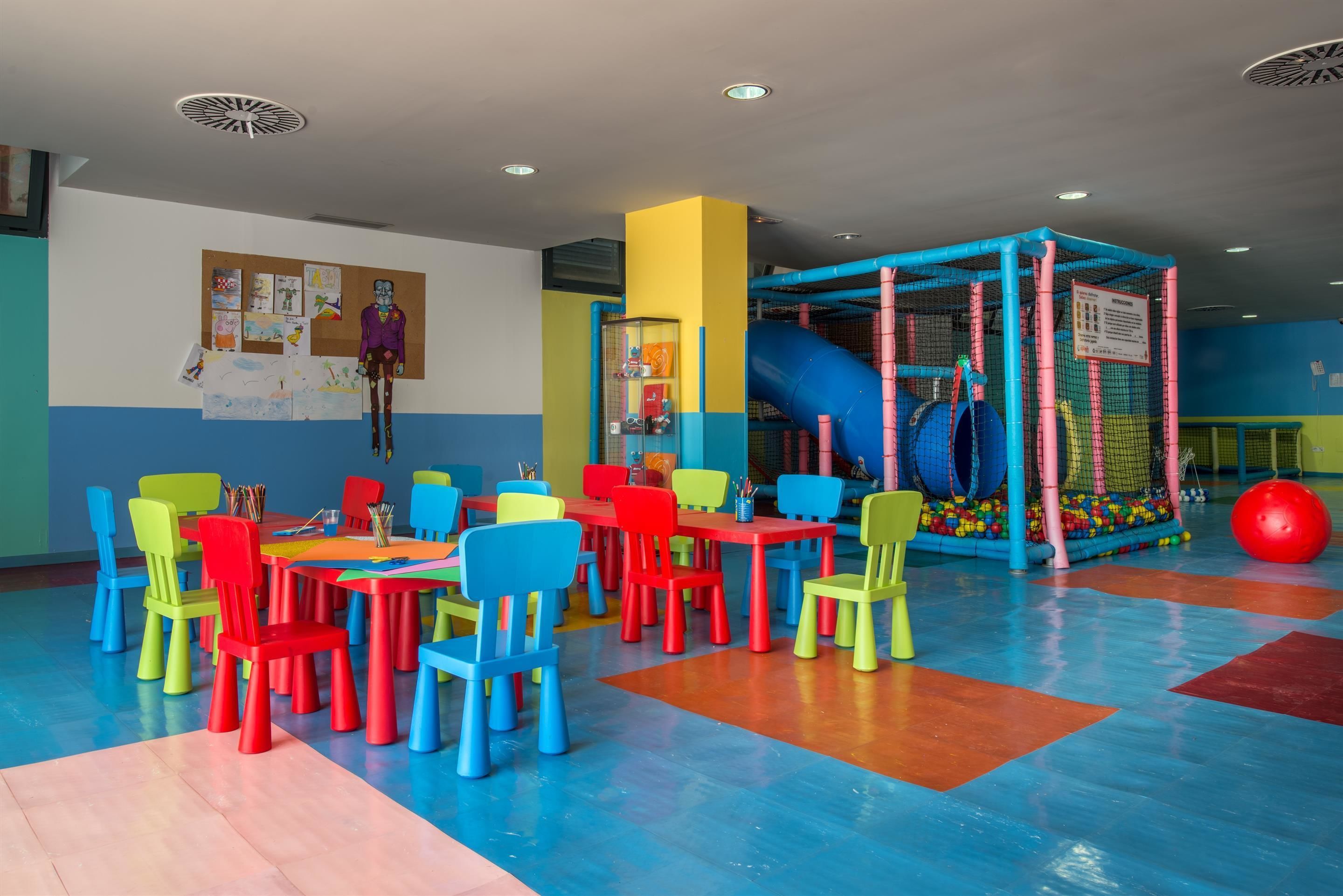 A children's play area with colorful tables and chairs, a play structure, and a bulletin board with drawings.