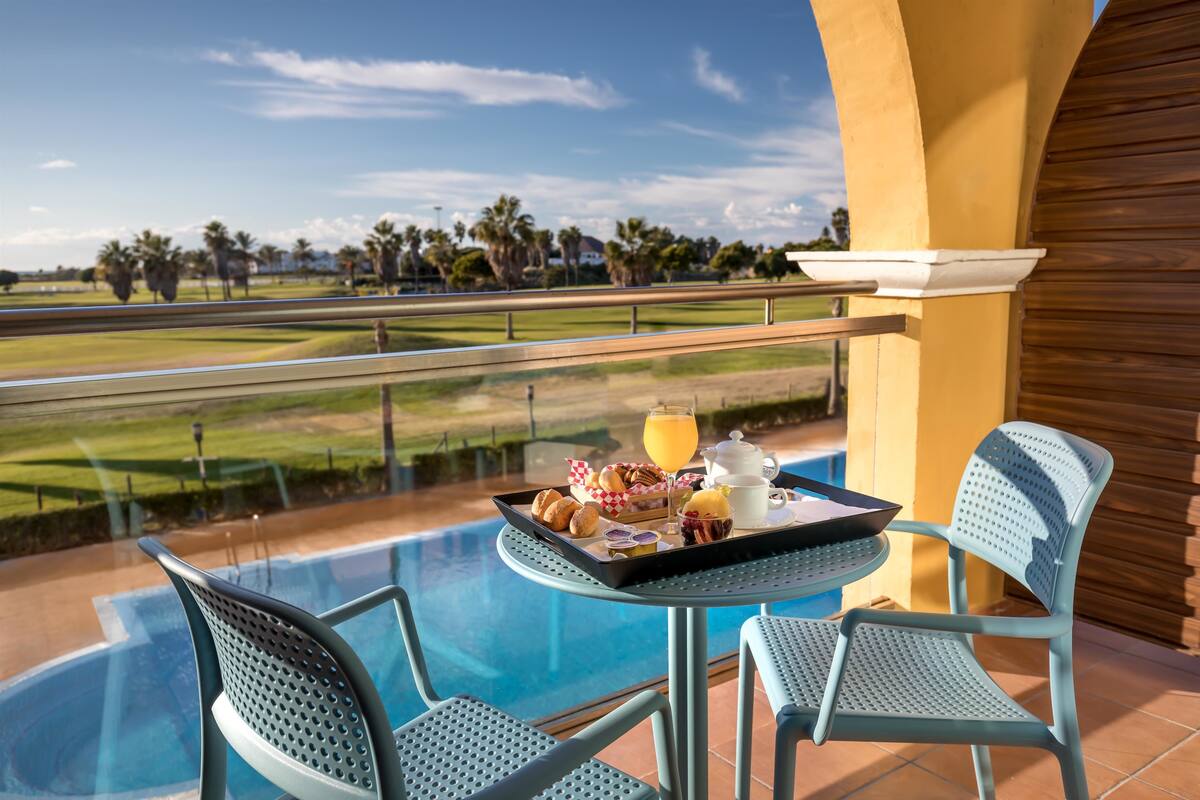 A balcony with a pool, two chairs, a table with breakfast items, and a view of a golf course.