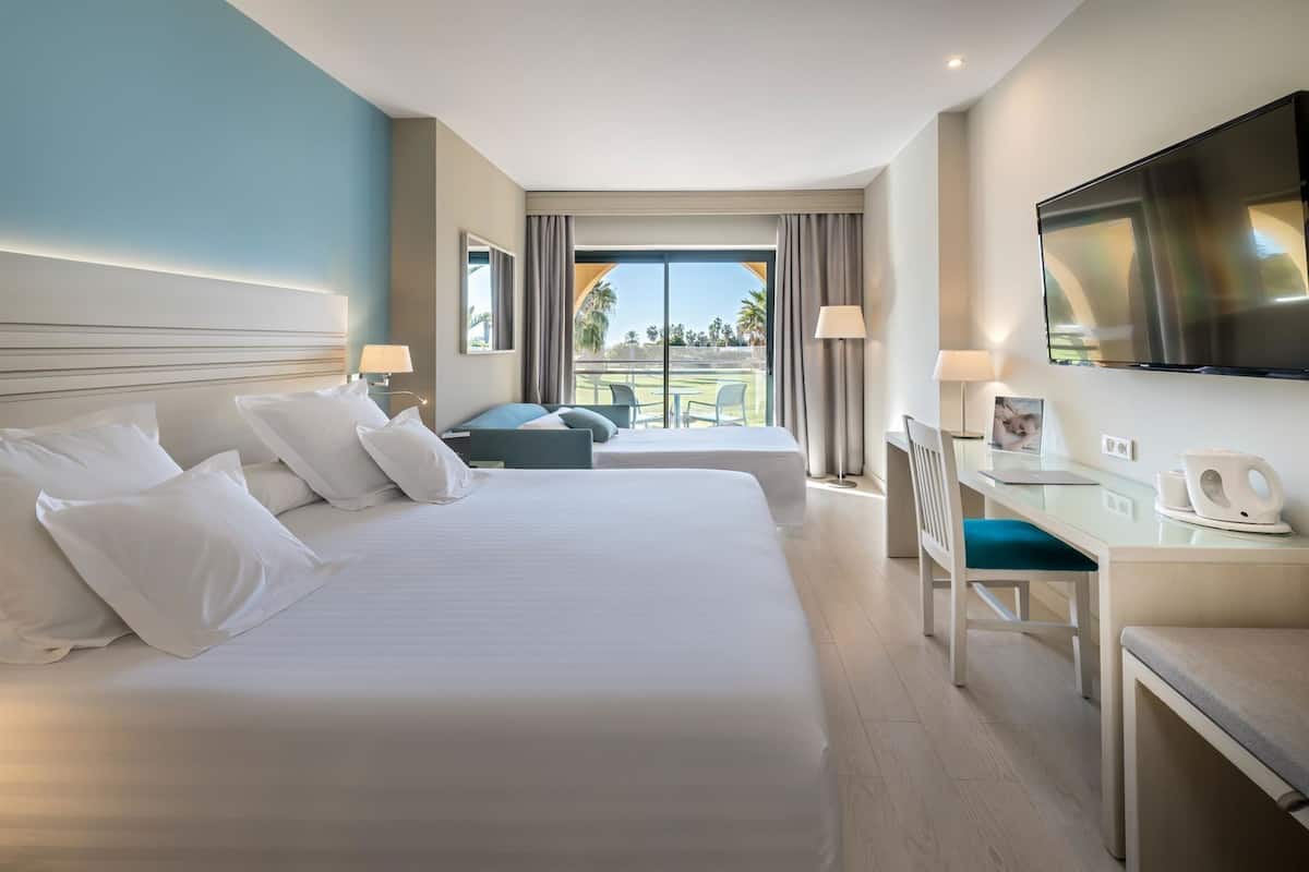 A hotel room with two beds, a desk, a chair, a TV, and a view of the outdoors.