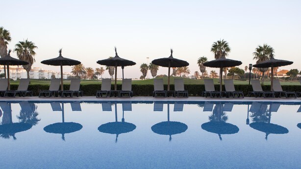 Una piscina con sillas de descanso y sombrillas, rodeada de palmeras y un cielo despejado.