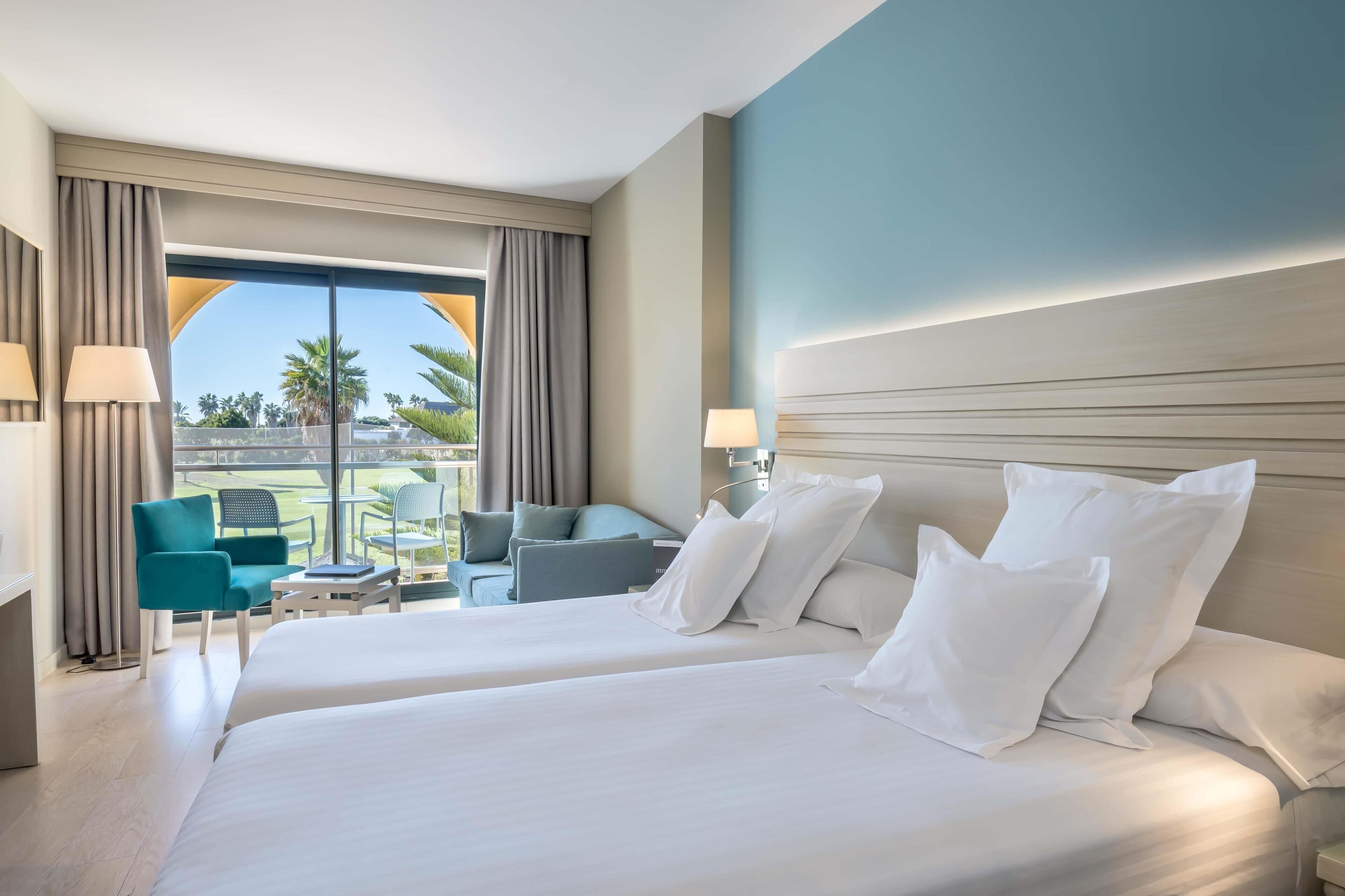 A hotel room with two beds, a sitting area with a table and chairs, and a large window with curtains.