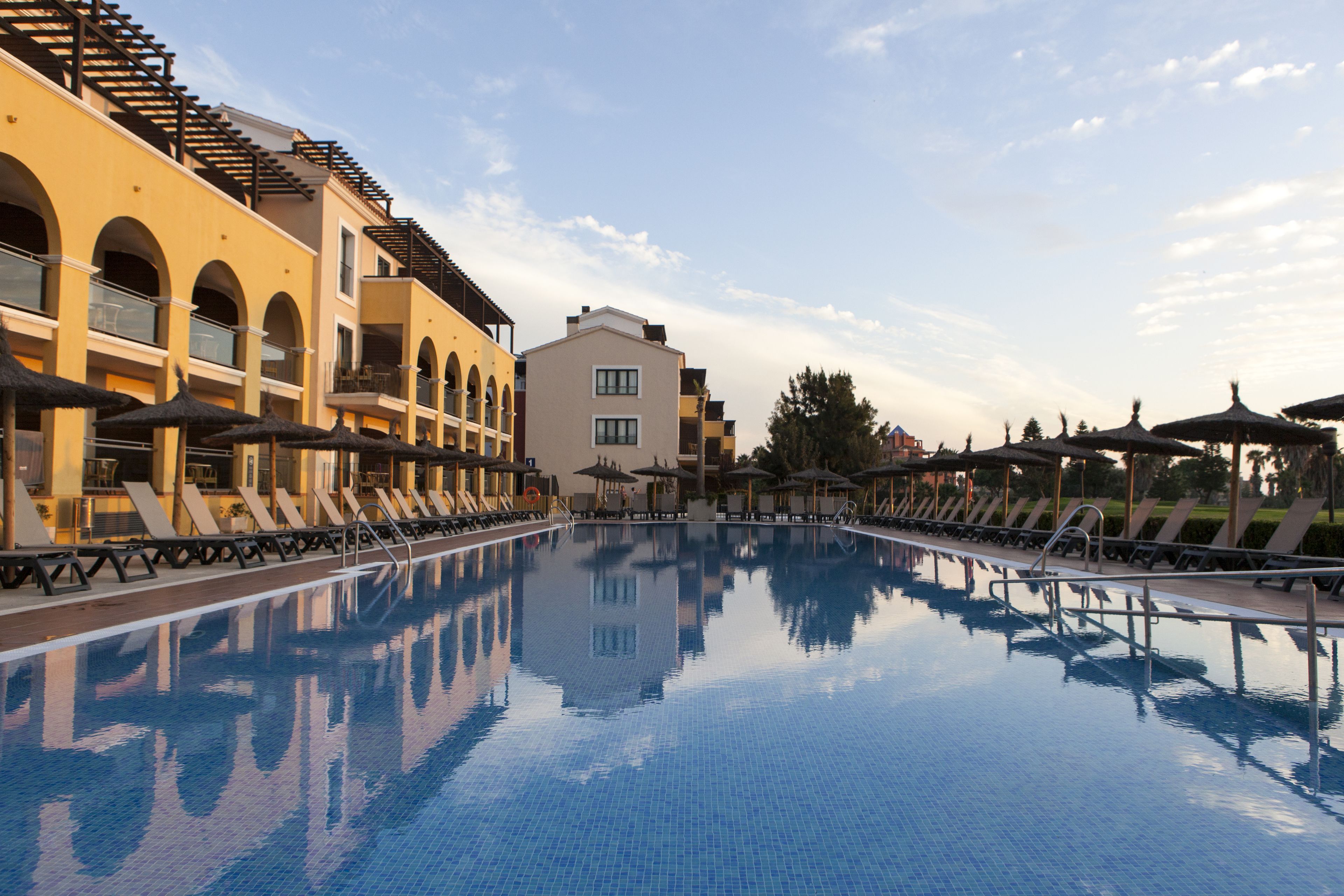 Une piscine entourée de bâtiments dotés de balcons et de fenêtres en arc, avec des chaises longues et des parasols à proximité.