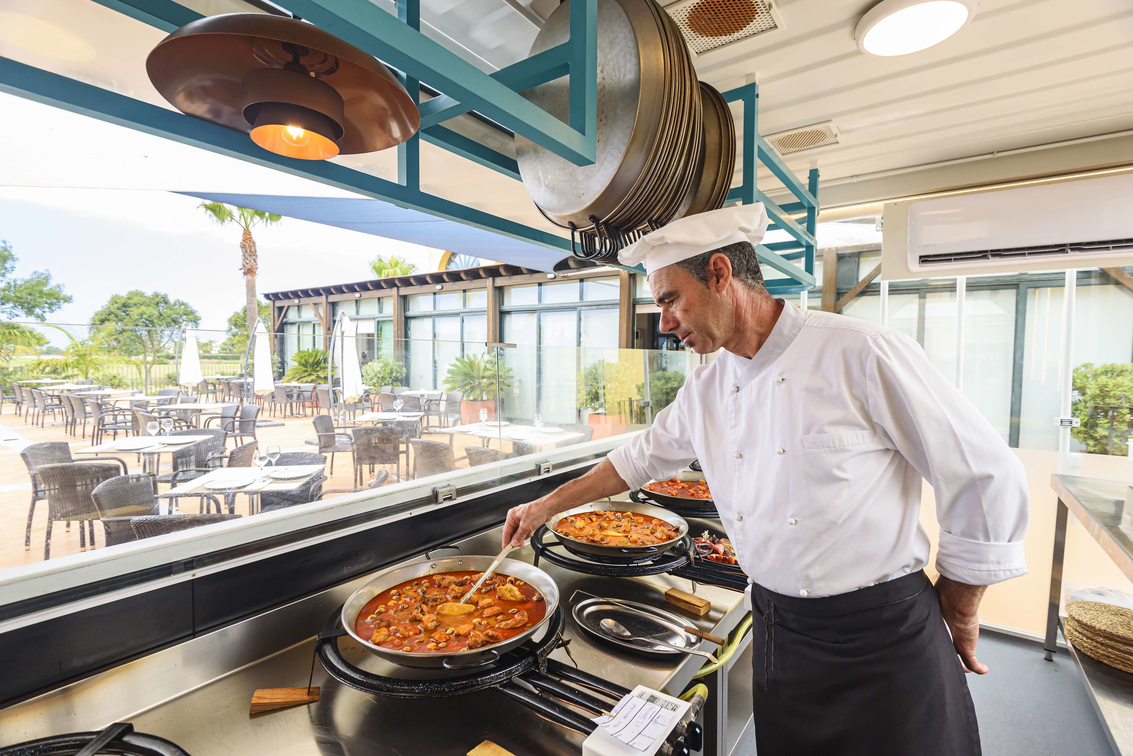 A chef preparing food in a kitchen with large pots and a view of outdoor seating.