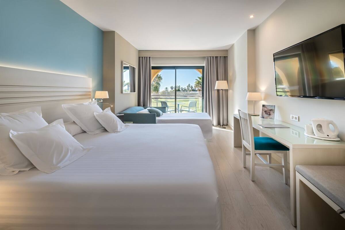 A hotel room with two beds, a desk, a chair, a TV, and a view of the outdoors.