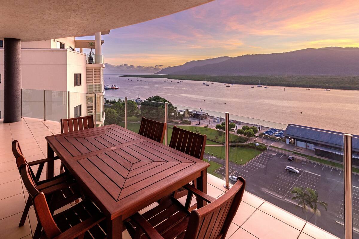 A balcony with a wooden table and chairs overlooking a body of water with boats and a bridge.