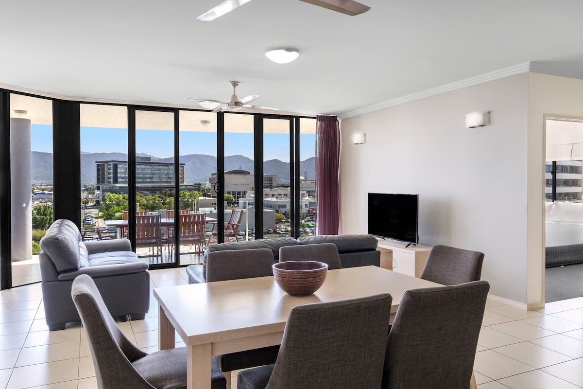A modern living room with a dining area, large windows offering a city view, a ceiling fan, and a flat-screen TV.