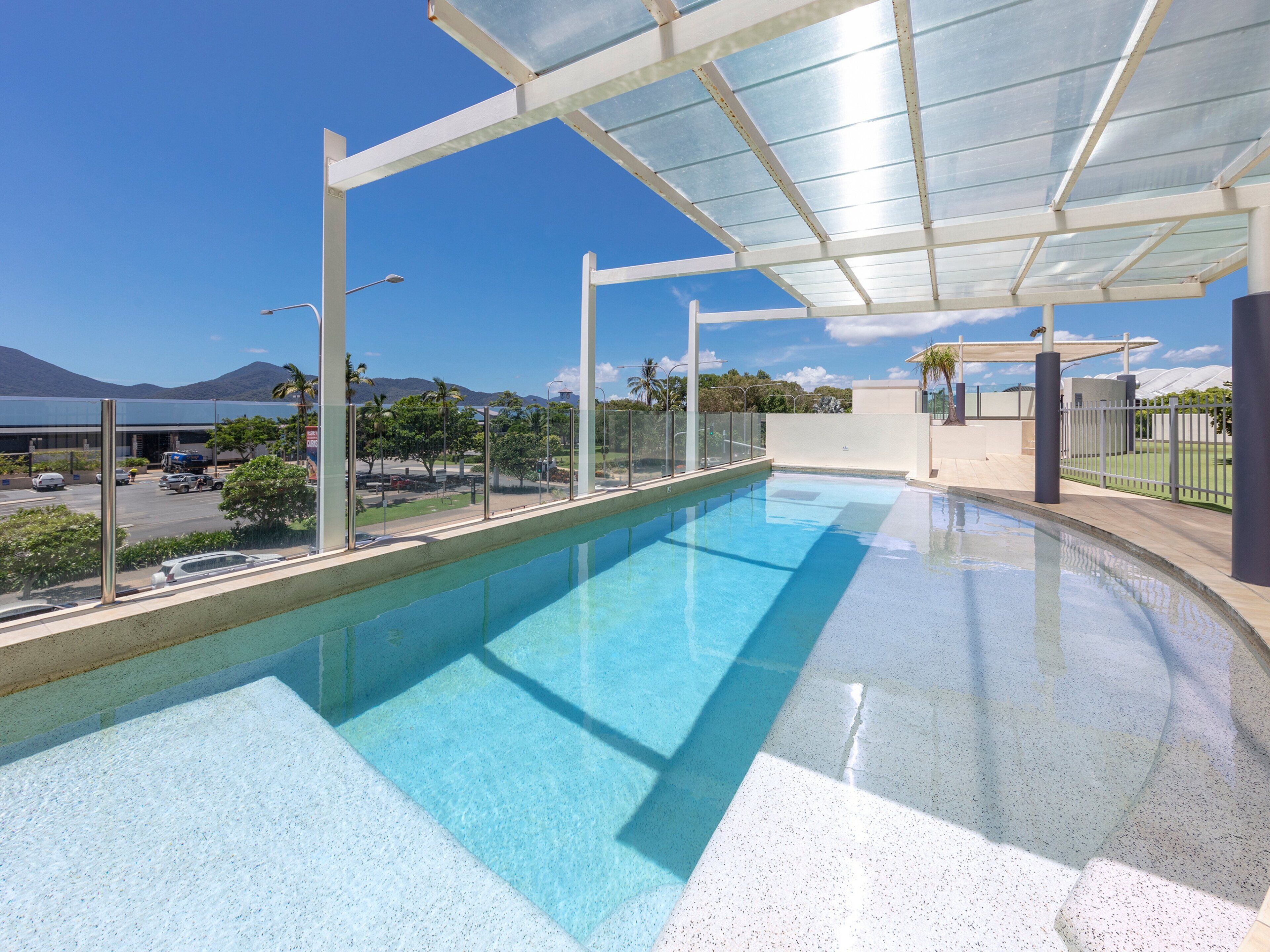A swimming pool with a glass enclosure, clear blue water, and a view of a parking lot and mountains in the background.