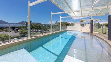 A swimming pool with a glass enclosure, clear blue water, and a view of a parking lot and mountains in the background.