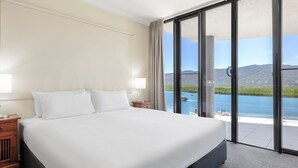 Een groot bed met witte lakens, een nachtkastje met een lamp, en uitzicht op een waterpartij door grote glazen deuren.