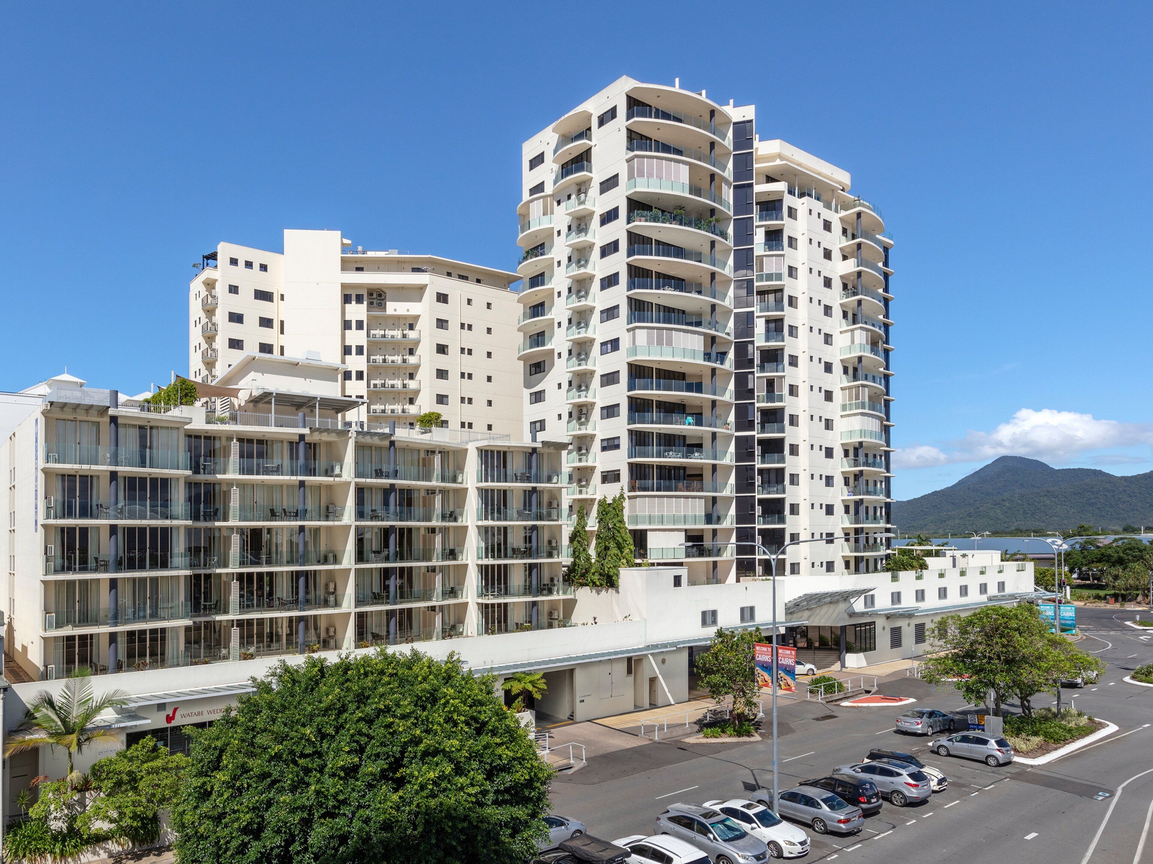 A modern multi-story hotel with balconies, a parking lot, and a mountain in the background.