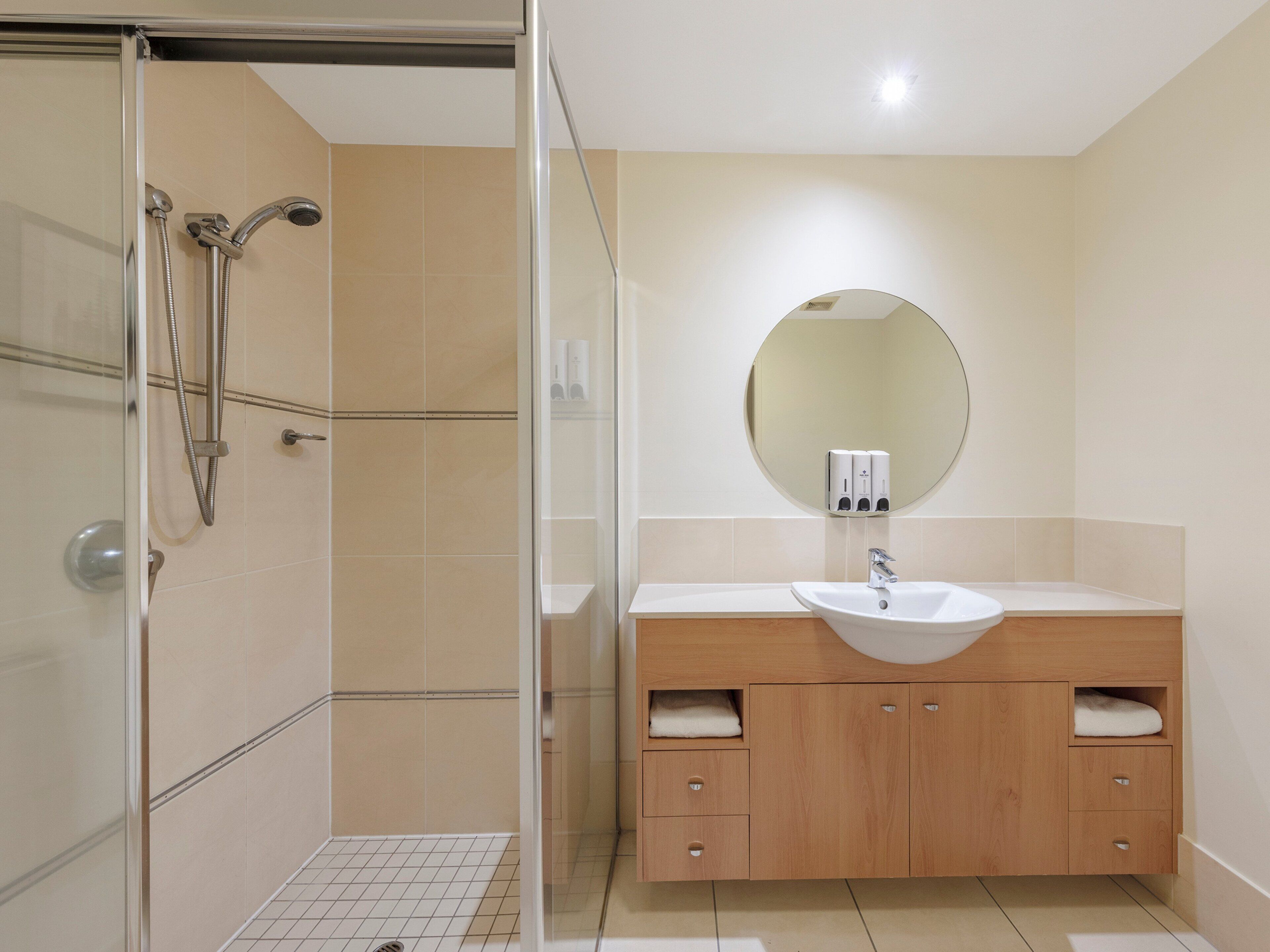 A modern bathroom with a glass shower enclosure, a white sink, wooden vanity, and a round mirror.