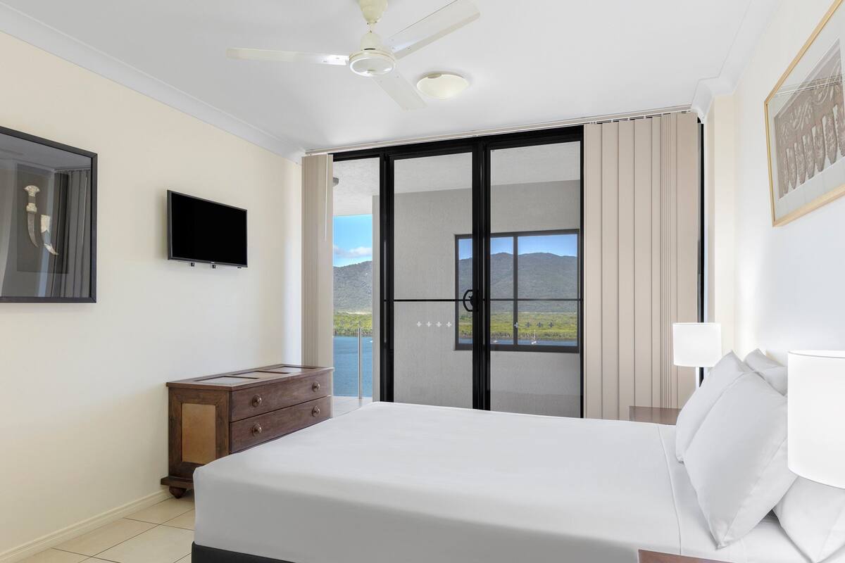 A bedroom with a bed, a dresser, a TV, and a balcony with a view of the mountains.