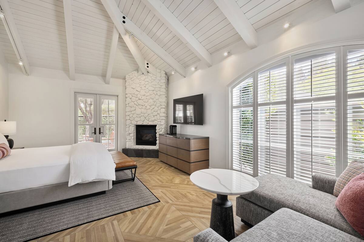 A modern bedroom with a large bed, a stone fireplace, a TV, and a view through large windows.