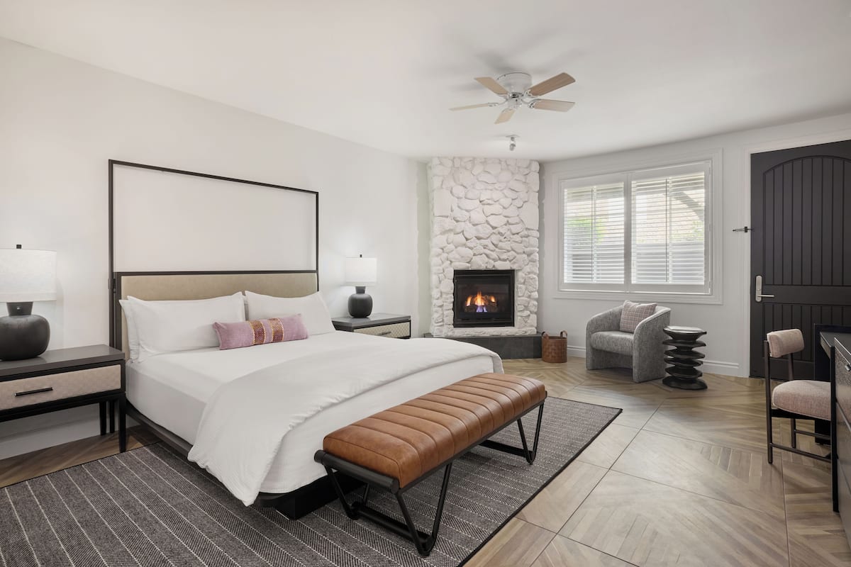 A modern bedroom with a large bed, a stone fireplace, a ceiling fan, and a view of the outdoors through the window.
