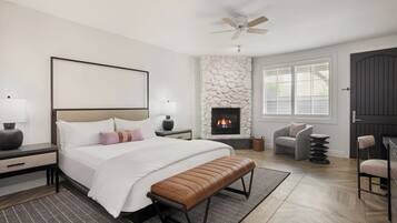 A modern bedroom with a large bed, a stone fireplace, a ceiling fan, and a view of the outdoors through the window.