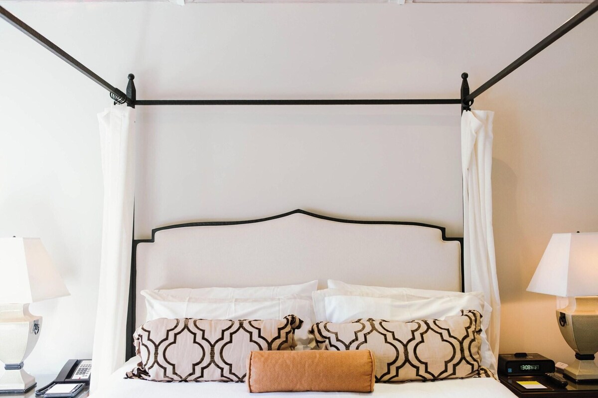 A hotel room with a four-poster bed, white bedding, patterned pillows, and a bedside table with a lamp and a phone.