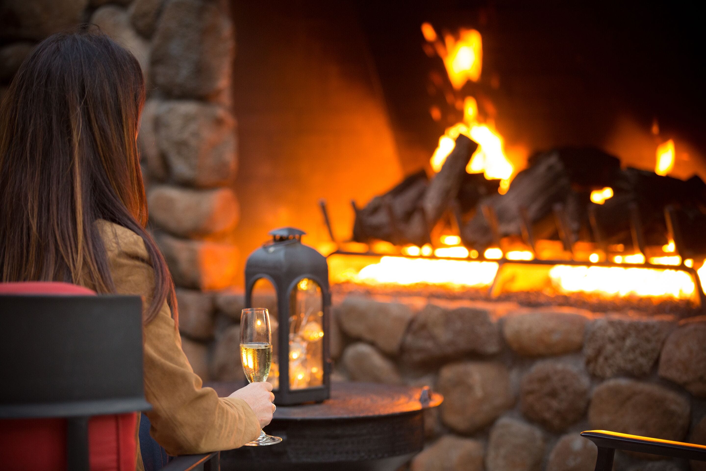 A person seated at a table with a glass of wine, facing a stone fireplace with a lantern.