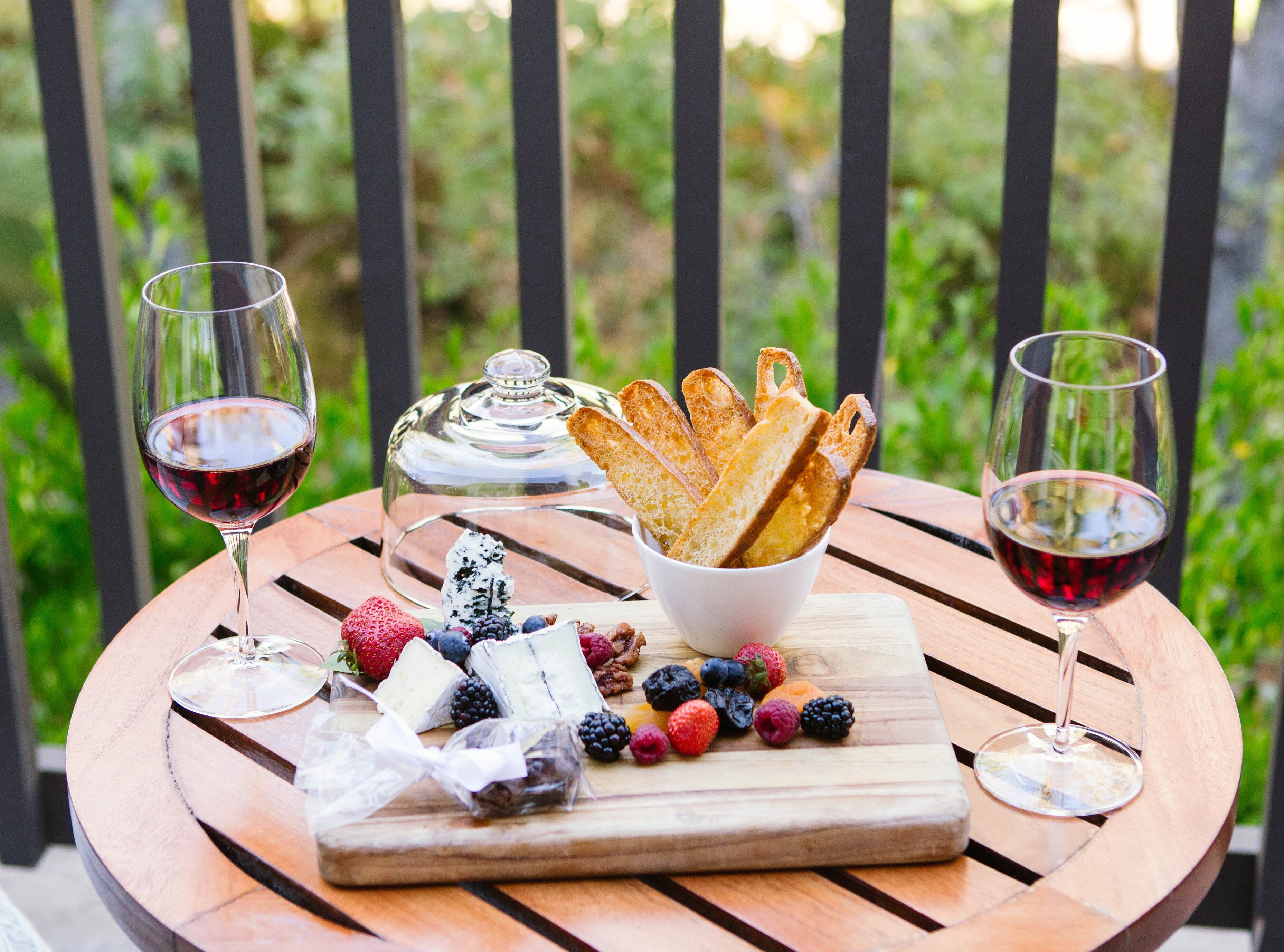 A wooden table with a cheese board, two glasses of red wine, and a glass dome container.